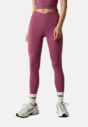 Eng anliegende pinke Leggings mit einem hohen, überkreuzten Bund, kombiniert mit weißen und schwarzen Sportschuhen und weißen Socken mit schwarzem Schriftzug.