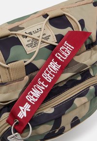 Sacca in tessuto mimetico con chiusura a zip, presenta un'etichetta rossa "RIMUOVERE PRIMA DEL VOLO" e un'etichetta beige con logo e dettagli testuali.