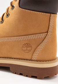 Hnědé kožené kotníkové boty s polstrovaným límcem, bílým prošíváním a vyraženým logem stromu na boku; gumová podrážka s texturovaným gripem.