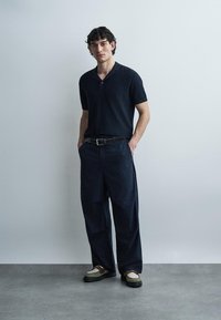 Fiatal férfi zsebre tett kézzel áll, fekete rövid ujjú galléros pólót, tengerészkék csíkozású bő nadrágot, fekete övet és két tónusú cipőt visel szürke padlón.