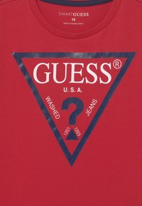 Rød bomull T-skjorte med en marineblå triangel-logo som har teksten "GUESS® U.S.A." og et spørsmålstegn, sammen med ordene "VASKET JEANS" og tall.