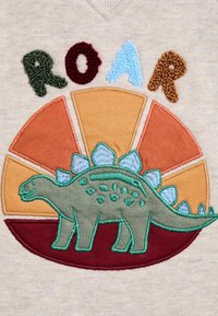 Camisa beige con un motivo de dinosaurio bordado. El dinosaurio es verde con acentos en azul y verde oscuro, situado sobre un diseño de resplandor multicolor.