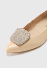 Chaussure plate beige à bout pointu avec une boucle carrée texturée en argent métallique ornée de petits accents scintillants. Surface lisse, design minimaliste.