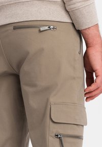 I pantaloni cargo beige presentano una tasca con zip anteriore, una tasca con patta laterale e una texture liscia. Un primo piano mette in evidenza i dettagli delle zip e delle cuciture.