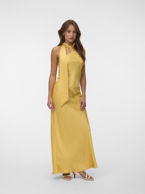 Femme portant une longue robe jaune à col montant avec un nœud au cou, debout devant un fond uni gris clair, portant des sandales beiges.