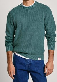 Grüner Strickpullover mit strukturiertem Muster, gerippten Bündchen und Saum. Über einem weißen T-Shirt getragen, kombiniert mit blauen Jeans.