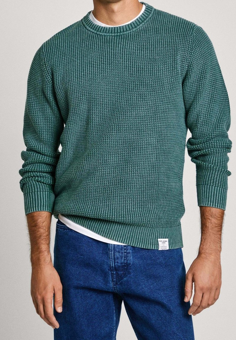 Grüner Strickpullover mit strukturiertem Muster, gerippten Bündchen und Saum. Über einem weißen T-Shirt getragen, kombiniert mit blauen Jeans.