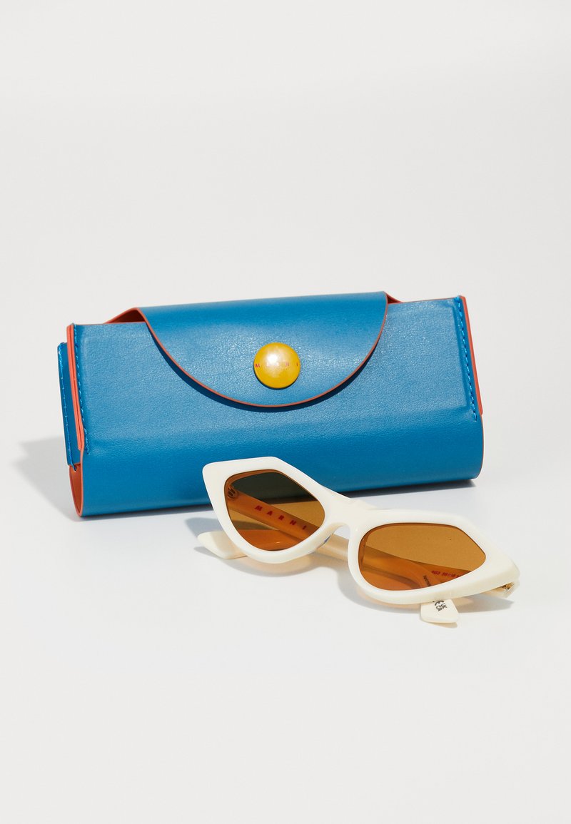 Occhiali da sole bianchi a forma di occhio di gatto con lenti marroni e una custodia in pelle blu con un bottone giallo. La custodia ha bordi rossi e un design a pattina curva.