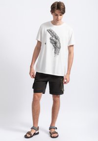 Witte katoenen t-shirt met een zwart geïllustreerd handontwerp, gecombineerd met zwarte cargoshorts en sandalen, staande tegen een lichte achtergrond.