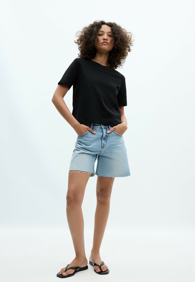 Femme aux cheveux bouclés portant un t-shirt noir à manches courtes, un short en jean clair et des sandales noires, debout les mains dans les poches.