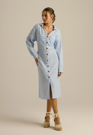 Jasnoniebieska sukienka koszulowa w paski, wykonana z bawełny, z długimi rękawami, zapięciem na guziki, kieszonką na piersi oraz bocznym rozcięciem, zestawiona z beżowymi espadrylami na koturnie.