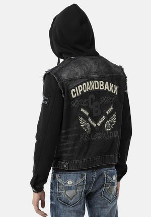 Mann trägt schwarze Kapuzenjacke mit "CIPOANDBAXX" und Flügelgrafiken auf dem Rücken, kombiniert mit blauen bestickten Jeans, von hinten gesehen.