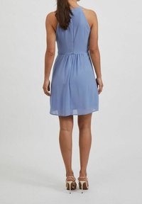 Robe bleu clair sans manches avec un col montant, une taille froncée et une jupe fluide tombant à mi-cuisse. Portée avec des sandales à talons hauts beige.