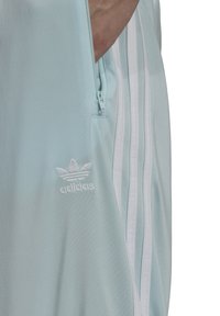 Svetle modré sportovní oblečení s kapsou na zip a třemi vertikálními bílými pruhy na boku. Vyšívané logo v bílé barvě.