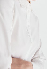 Chemise blanche à manches longues en tissu lisse, avec un col classique et des boutons légèrement brillants, présentée partiellement sur une silhouette.