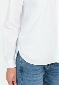 Chemise blanche à manches longues en tissu doux, avec un ourlet arrondi et un détail logo circulaire subtil en bas à gauche.