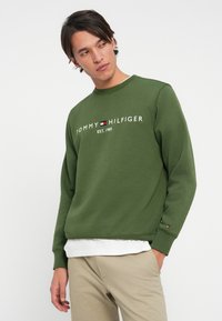 Olive green sweatshirt with crew neck, featuring white "TOMMY HILFIGER" logo and red, white, and blue emblem on the sleeve; cotton blend material.