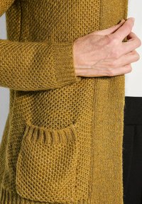 Senfgelbe gestrickte Strickjacke mit strukturiertem Muster, ausgestattet mit einer einzigen Vordertasche und gerippten Bündchen. Weiches Material und offenes Vorderdesign.