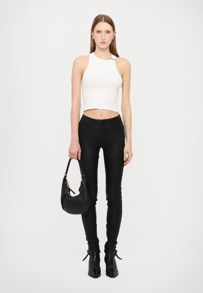 Bílý zkrácený top bez rukávů, černé skinny džíny a černé kotníkové boty. Modelka drží černou kabelku s křivkovým designem a viditelným prošíváním.