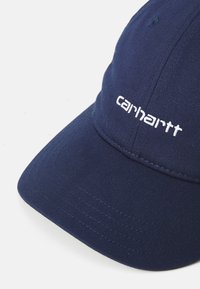 Cappellino blu navy con visiera arrotondata. Presenta il logo "carhartt" ricamato in bianco, una texture a coste e cuciture a punto.