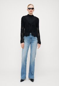 Cardigã preto de crochê com design de malha aberta, fechamento com laço na cintura, combinado com jeans azuis claros de boca de sino e sapatos de salto pretos de bico.