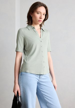 Woman wearing light green short-sleeve buttoned shirt and light blue jeans, holding a black textured handbag against a plain background.