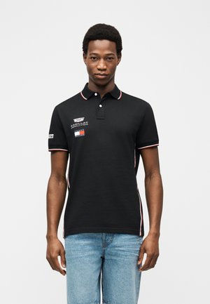 Homme portant un polo noir de l'équipe Cadillac Formula E avec des logos et un jean bleu clair, debout devant un fond blanc uni.