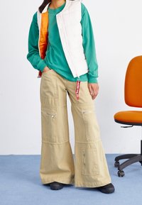 Grüner Sweatshirt, weiße gesteppte Weste mit orangefarbener Fütterung und beigen, weiten Cargo-Hosen mit Reißverschlüssen; schwarze Schuhe. Schlichter Hintergrund.
