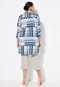 Langer, gemusterter Mantel in Blau und Weiß mit kreisförmigen Designs. Kombiniert mit beigefarbenen, knöchellangen Hosen und Absatzsandalen. Glatte Stoffstruktur.