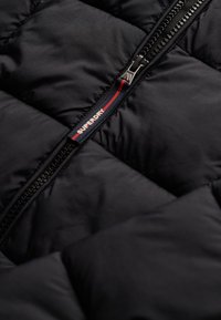 Veste matelassée noire dotée d'une fermeture éclair visible avec tirette de marque, tissu texturé et design matelassé, mettant l'accent sur la chaleur et la fonctionnalité.
