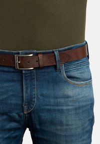 Brun læderbælte med en sølvmetalkrog, båret over blå denimjeans med let slid og kontrastsyet detaljer.