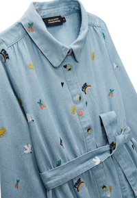 Chemise en denim bleu clair avec un col, ornée de motifs colorés brodés de soleils, d'arcs-en-ciel et d'oiseaux. Comprend un détail de taille nouée.