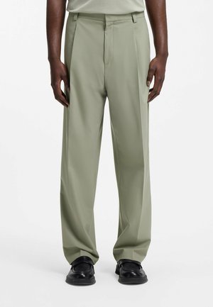 Mand iført lysegrønne vidde bukser og sorte lædersko, stående mod en ensfarvet hvid baggrund.