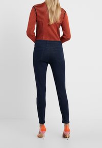 Jeans oscuros ajustados de cintura alta, con bolsillos traseros y una textura suave, combinados con zapatos de tacón alto en naranja y rosa.