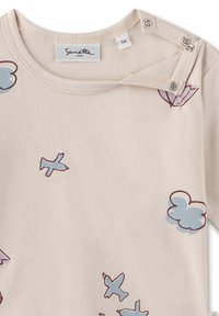 Helles beigefarbenes Baumwoll-T-Shirt mit blauen und lila Grafiken von Flugzeugen und Wolken. Mit Druckknopfverschlüssen an der Schulter für ein einfaches Anziehen.