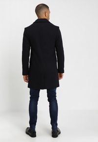 Abrigo largo de mezcla de lana negra con solapa de muesca, que presenta un corte entallado, textura suave y una abertura trasera sencilla. Lleva puesto sobre unos jeans azules.