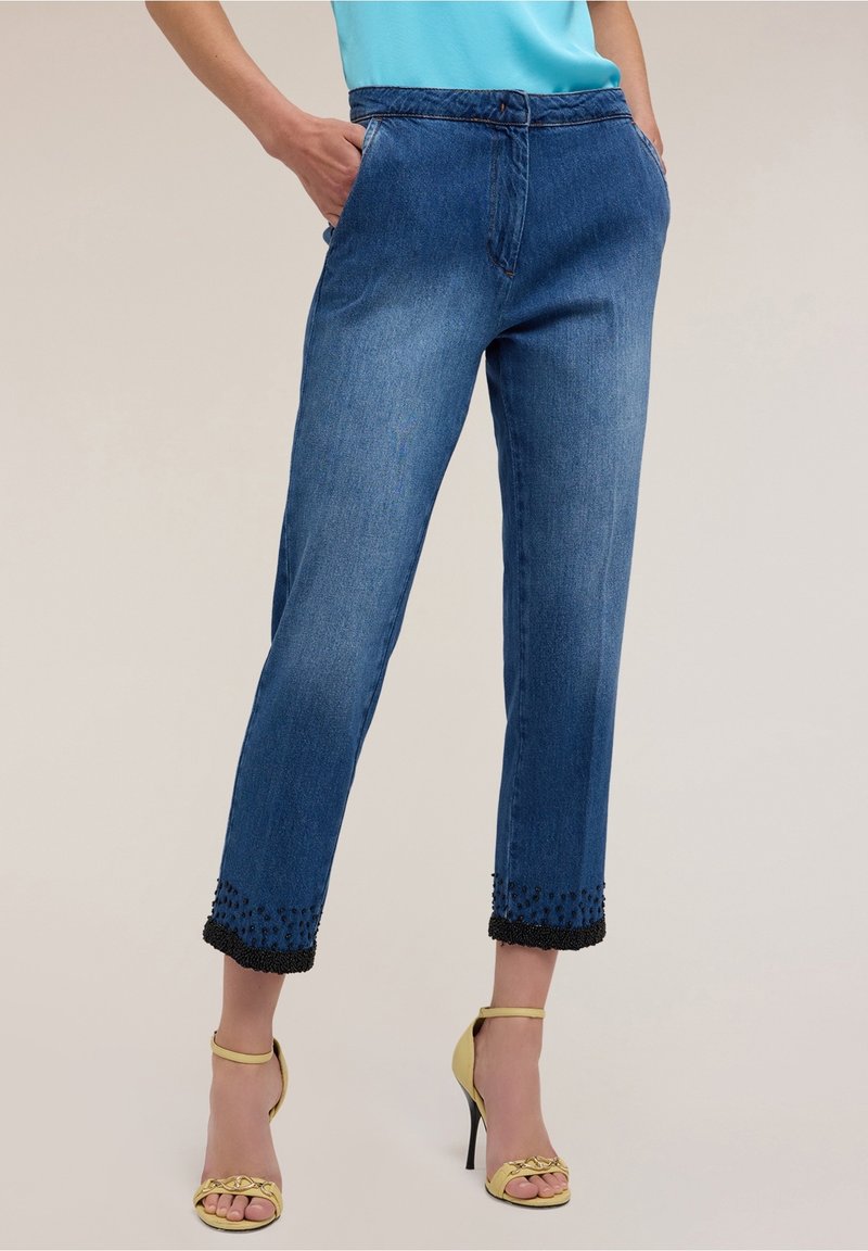 Woman wearing cropped blue jeans with black beaded hems and yellow high-heeled sandals, hands in pockets, light blue top visible.