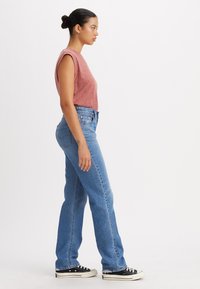 Rood gemêleerde mouwloze top gecombineerd met hoogwaist blauwe jeans, met een relaxte pasvorm en klassieke denimtextuur. Zwarte sneakers maken de look compleet.