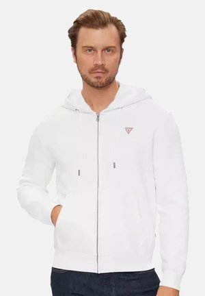 Man met witte hoodie met ritssluiting en klein driehoekig logo, staand met één hand in de zak tegen een effen achtergrond.