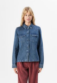 Blonde woman wearing a buttoned blue denim jacket with chest pockets and rust-colored corduroy pants, standing against a white background.