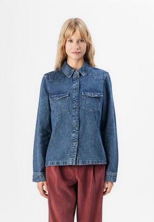 Blonde woman wearing a buttoned blue denim jacket with chest pockets and rust-colored corduroy pants, standing against a white background.