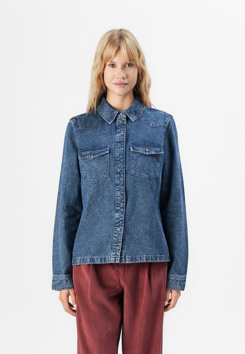 Blonde woman wearing a buttoned blue denim jacket with chest pockets and rust-colored corduroy pants, standing against a white background.