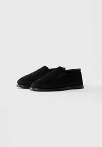 Une paire de chaussures slip-on en suede noir avec des détails brodés le long de la semelle, présentée sur un fond clair uni.