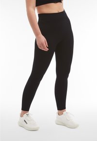 Leggings neri con una vita alta e una texture liscia, abbinati a scarpe sportive bianche con dettagli testurizzati e chiusure con lacci.