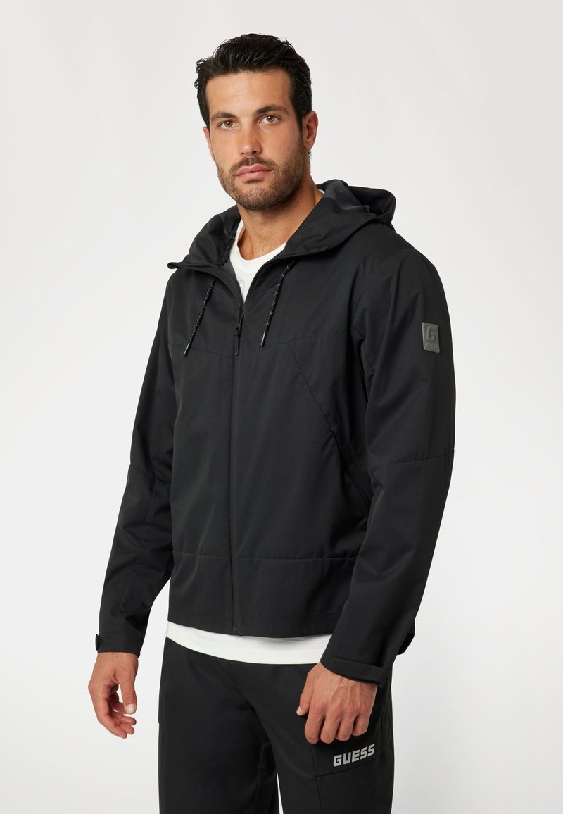 Juoda užtrauktuku užsegama striukė su kapišonu, pagaminta iš glotnaus, lengvo audinio. Yra raišteliai, logotipas ant rankovės ir elastingi rankogaliai.