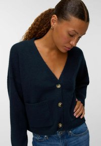 Sieviete ar zeltainām riņķa auskarām, tērpusies tumši zils pogājams džemperis ar kabatām un zila džinsos, skatās uz leju, roka uz gurniem.