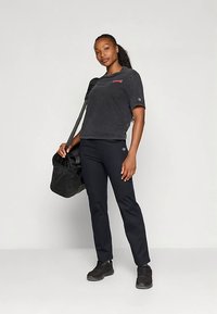 Ženska v črni vsakdanji oblačili, ki drži črno športno torbo čez ramo, stoji ob enobarvni svetli ozadju.