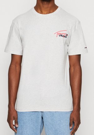 Homme portant un t-shirt gris clair Tommy Jeans avec un petit logo sur la poitrine et la manche, associé à un jean en denim bleu clair.