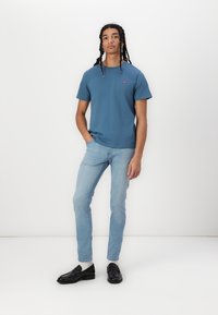 Kék pamut póló rövid ujjakkal, világoskék slim-fit farmernadrággal párosítva. Fekete loaferek és fehér zoknik egészítik ki az öltözéket.