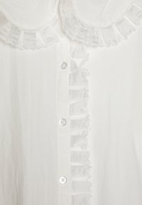 Hvit bluse med rynket krage og frontplacket. Laget av lett stoff, med dekorative volanger og knapper.